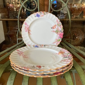 May include: A set of three white porcelain plates with a floral design. The plates have a scalloped edge and a gold trim. The flowers are pink, purple, and blue.