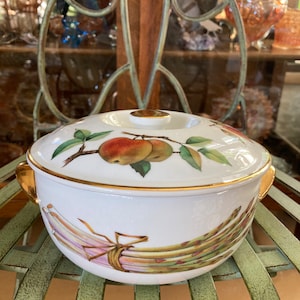 Peut inclure: Plat à gratin en porcelaine blanche avec un bord doré et un couvercle. Le plat présente un motif peint à la main d'asperges et de pommes sur une vigne verte.