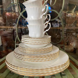 May include: A collection of white teacups and saucers with gold edging. The teacups are stacked on the saucers, forming a tiered arrangement. The rims of the cups and plates are adorned with a gold border. The set is displayed on a green metal surface.