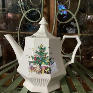 May include: White porcelain teapot with a Christmas tree design. The teapot features a geometric shape with a pointed lid and a handle. The Christmas tree is decorated with ornaments and presents. The teapot is sitting on a green metal surface.