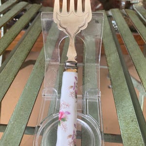 May include: A silver fork with a white porcelain handle decorated with pink and purple flowers. The handle has a floral design.