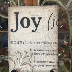 May include: A decorative square plaque with the word "Joy" in large black letters. The plaque includes a definition of "Joy" and a decorative floral design. The background has a crackled, aged appearance.