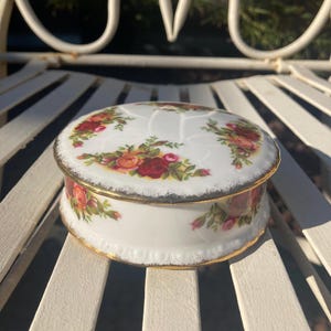 May include: A round white porcelain trinket box featuring a colourful floral pattern of red, orange, and yellow roses with green leaves. The lid and base are accented with a worn gold-toned trim along the edges, and the surface has a subtle embossed texture.