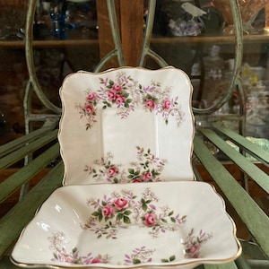 May include: Two square, white porcelain dishes with a gold rim and a floral pattern of pink roses, green leaves, and purple sprigs. The dishes are stacked on top of each other.