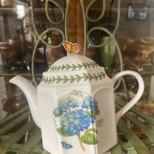 May include: White ceramic teapot with a butterfly on the lid. The teapot features a blue hydrangea floral design and green leaf accents. The teapot is sitting on a green metal surface.