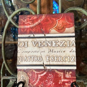 May include: A red and white ceramic tile with a cracked surface. The tile features a decorative design and the text "DI VENEZIA Composta in Musica da QUATTRO ESERCIZI".