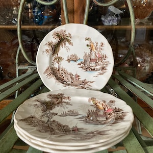 May include: Set of four brown and white ceramic plates featuring a cottage scene design. The plates have a scalloped edge and are stacked on top of each other.