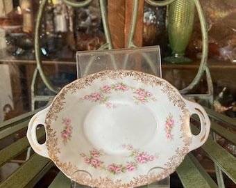May include: An oval porcelain dish with two side handles, featuring a white base, a delicate pattern of small pink roses, and ornate gold filigree trim along the scalloped edges. The dish is displayed on a clear stand for visibility.