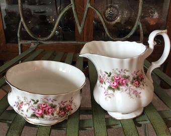 May include: A white porcelain creamer and sugar bowl set with a floral pattern of pink roses and green leaves. The set is sitting on a green metal chair.