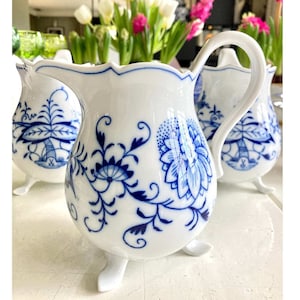 May include: Three white porcelain cream jugs with blue floral designs and three legs. The jugs have a scalloped edge and a handle.