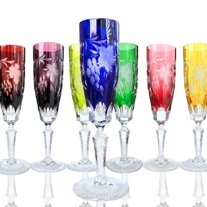 Könnte beinhalten: Set mit sieben farbigen Sektgläsern. Jedes Glas hat eine andere Farbe: Rot, Lila, Gelb, Blau, Grün und Rosa. Die Gläser haben klare Stiele und Böden und sind mit geätzten Trauben- und Blattmotiven verziert.