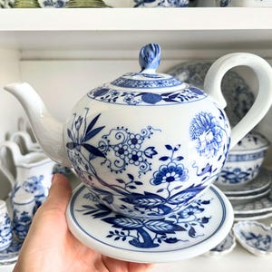 Op de afbeelding: Een witte theepot met een blauw en wit bloemenpatroon. De theepot heeft een deksel en een handvat. De theepot staat op een witte schotel met een blauw en wit bloemenpatroon.