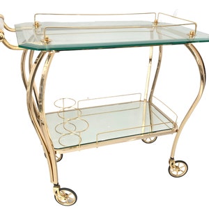 May include: A gold-coloured drinks trolley with two glass shelves and a handle with a white accent. The cart has a curved frame and small wheels. It features a built-in wine glass holder.