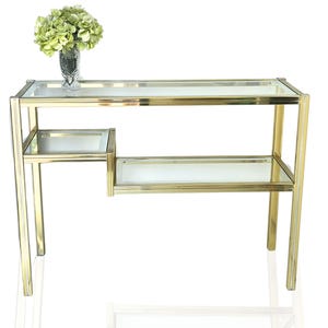 Peut inclure: Une console dorée avec deux étagères en verre et une petite étagère latérale. La table est de forme rectangulaire et est ornée d'un vase de fleurs vertes. La table est en métal et en verre.