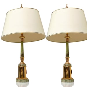 Op de afbeelding: Twee bijpassende tafellampen met crèmekleurige kappen en gouden details. De lampen hebben een groene middenkolom en een decoratieve gouden voet. Deze lampen zijn ontworpen voor gebruik binnenshuis.