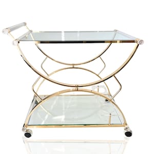 May include: A gold-toned bar cart with two clear glass shelves and a curved frame design. The cart has clear acrylic handles and sits on small rolling wheels. The overall design is elegant and functional, suitable for serving drinks and displaying items.