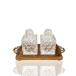 Op de afbeelding: Een vintage zout- en peperstel. Twee heldere, diamantpatroon glazen shakers met heldere doppen staan op een goudkleurige metalen dienblad met sierlijke handgrepen. De set is ontworpen voor gebruik op tafel.