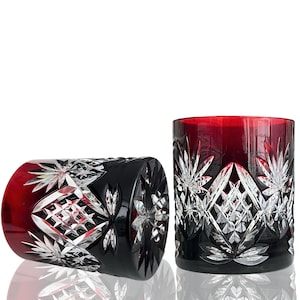Op de afbeelding: Twee rode en zwarte kristallen whiskyglazen met ingewikkelde patronen van sterren en geometrische sneden. Eén glas staat rechtop, de andere ligt op zijn kant. De glazen staan op een witte ondergrond.