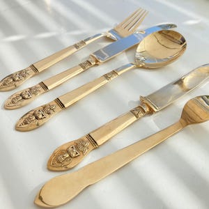 Op de afbeelding: Een set van vijf vergulde bestekstukken, waaronder een vork, een mes, een lepel en twee extra vorken. Het bestek heeft ingewikkelde, sierlijke ontwerpen.