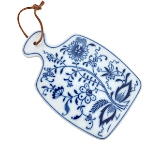 Op de afbeelding: Een wit porseleinen snijplank met een blauw bloemenpatroon en een bruine leren lus om op te hangen.