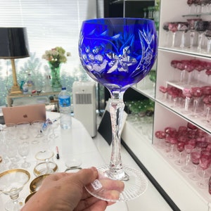 Puede incluir: Una copa de vino de cristal azul cobalto con un diseño cortado. La copa tiene un tallo largo y una copa ancha. La copa está siendo sostenida por una mano.