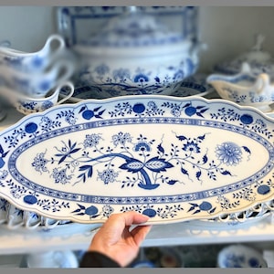 Op de afbeelding: Een ovale serveerschaal met een blauw bloemmotief op een witte achtergrond. De schaal heeft een geschulpte rand en maakt deel uit van een servies. Andere onderdelen van de set, zoals een sauskom en een terrine, zijn zichtbaar op de achtergrond.