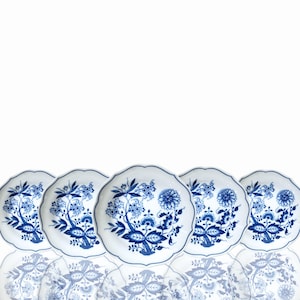 Puede incluir: Cinco platos de cerámica blanca con borde festoneado, cada uno adornado con un diseño floral azul. Los platos están dispuestos en fila, con sus reflejos visibles debajo. El patrón floral incluye flores y hojas estilizadas.