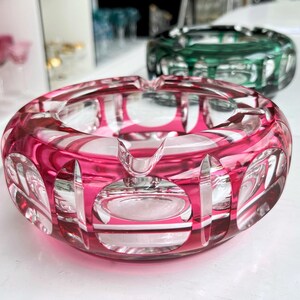 Op de afbeelding: Close-up van een roze kristallen asbak met een cirkelvormig ontwerp. De asbak heeft een reeks ovale uitsparingen rond de rand en een gebogen rustplaats voor een sigaret. Op de achtergrond is een groene asbak zichtbaar.