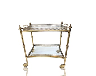 May include: A vintage gold-toned bar cart with two glass shelves and decorative metal detailing. The cart has a rectangular top shelf with handles and a lower shelf. It stands on four small wheels for easy mobility.