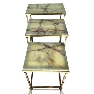 Puede incluir: Un juego de tres mesas nido con tapas cuadradas de ónix verde claro con vetas marrones. Las mesas tienen una estructura metálica de color latón. Las mesas están dispuestas en una configuración apilada.