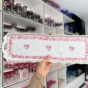 Op de afbeelding: Een rechthoekige witte keramische serveerschaal met een roze bloemenontwerp. De schaal heeft een geschulpte rand en gouden rand. Roze rozen zijn rond de rand en in het midden gerangschikt. De schaal heeft twee handvatten.