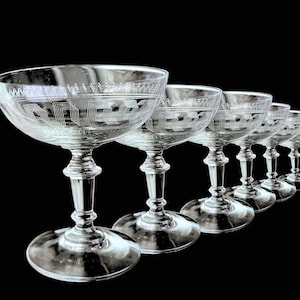 Könnte beinhalten: Sechs klare Champagner-Coupe-Gläser mit geätzten geometrischen Mustern. Die Gläser haben eine breite, flache Schale, einen detaillierten Stiel und einen runden Fuß. Die Gläser sind diagonal vor einem schwarzen Hintergrund angeordnet.