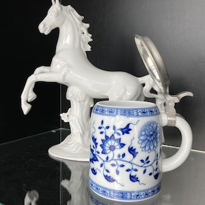 Op de afbeelding: Een wit porseleinen paardenbeeldje met een blauw-wit porseleinen bierkan met een tinnen deksel.