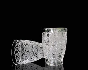 Op de afbeelding: Twee heldere glazen bekers met een ingewikkeld wit geëtst ontwerp. De bekers zijn leeg en staan op een zwart oppervlak.