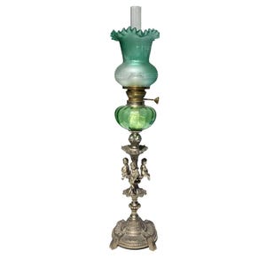 Op de afbeelding: Een antieke olielamp met een groene glazen kap en een decoratieve metalen voet. De lamp heeft een gerimpelde bovenkant, een ronde groene glazen reservoir en een gedetailleerde voet met kleine figuren.