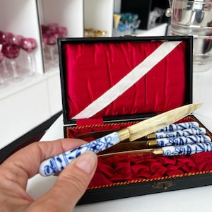 Op de afbeelding: Een set van vier kleine messen met gouden lemmeten en blauw-witte porseleinen handvatten, gepresenteerd in een zwarte doos met een rode fluwelen binnenkant. De handvatten hebben een bloemmotief.