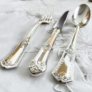 Op de afbeelding: Een set van drie zilverkleurige metalen bestekstukken bestaande uit een vork, een mes en een lepel. Elk stuk is voorzien van een sierlijk, reliëf decoratief patroon op het handvat, inclusief schelpvormige motieven en ingewikkeld krulwerk.