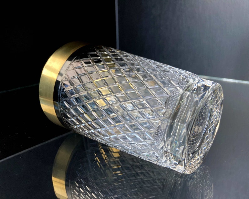 May include: A clear glass tumbler with a gold rim and a diamond pattern. The glass is empty and is sitting on a black surface.