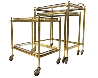 May include: A set of three gold-tone metal serving carts with glass shelves and wheels. The carts are nested together, with the largest cart on the bottom and the smallest cart on top.