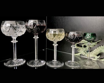 May include: Five stemmed crystal glasses with intricate cut designs. The glasses are filled with different colored liquids, including red, white, and green.