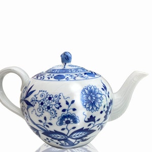 Peut inclure: Une théière blanche avec un motif floral bleu et blanc. La théière a un corps rond et un bec verseur sur le côté droit. La poignée est sur le côté gauche.