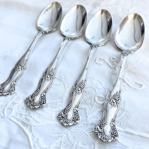 May include: Four antique silver spoons with ornate grape and leaf designs on the handles. The spoons are arranged on a white, textured surface, reflecting light. The spoons are likely for serving or eating.