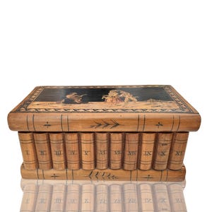 Puede incluir: Caja de rompecabezas de madera diseñada para parecerse a una pila de libros. La parte superior presenta un panel negro con una escena pintada, y los lados están tallados para parecerse a lomos de libros con números romanos.