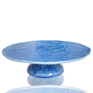 May include: A blue ceramic cake stand with a floral embossed design. The stand has a round, raised base and a wide, flat top. The stand is a light blue colour and is suitable for displaying cakes and other desserts.
