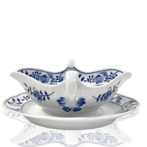 Op de afbeelding: Een witte porseleinen sauskom met een blauw bloemenmotief en een bijpassend schoteltje. De kom heeft een handvat aan de ene kant en een tuit aan de andere.