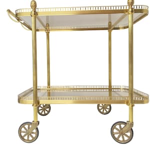 May include: A gold-toned metal serving cart with two glass shelves and four wheels. The cart has a decorative design on the top and bottom edges of the shelves.