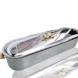 May include: A silver-toned, oblong serving dish with a gold-colored fish accent. The dish has a rounded shape and a small, decorative handle on one end. The interior is reflective, and the overall design is sleek and modern.