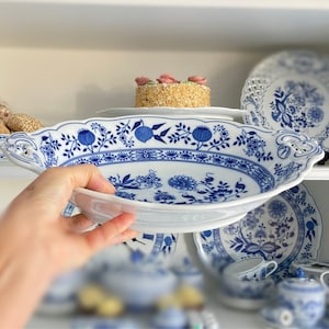 Op de afbeelding: Een ovale serveerschaal van wit porselein met een blauw bloemmotief. De schaal heeft handvatten en een geschulpte rand. Andere bijpassende blauw-witte serviesdelen, zoals borden en kopjes, zijn zichtbaar op de achtergrond.
