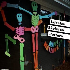 May include: Two large, colorful, articulated wooden skeleton decorations hanging vertically. The skeletons are painted in bright shades of green, blue, pink, and orange. A banner with the text "Lifesize Skeleton Pattern" and "Happy Halloween" is visible.
