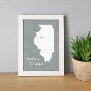 May include: A framed print featuring the outline of the state of Illinois in white, with a small purple heart marking a location. The background is a textured gray, and the words "your town" are written in cursive. The frame is white.